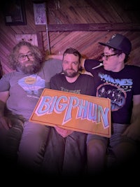 three men sitting on a couch holding a big phun sign